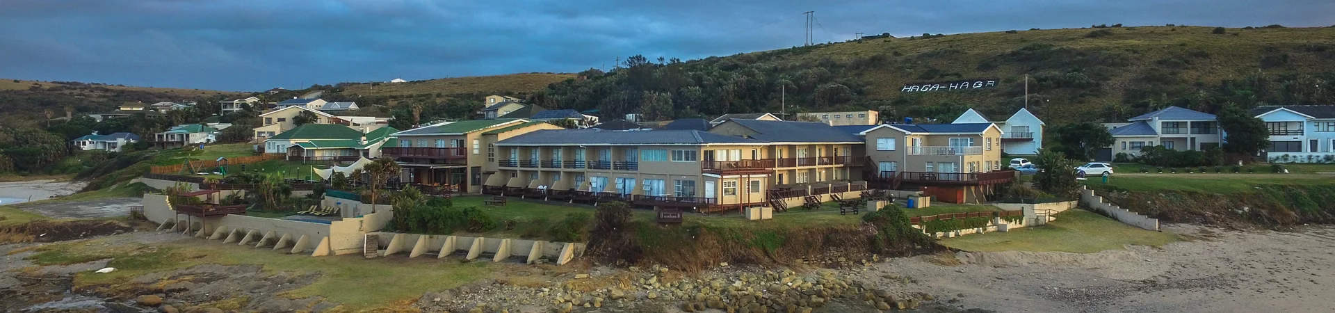A seaside hotel and several houses sit on a grassy hill overlooking rocky shores and waves under a cloudy sky.
